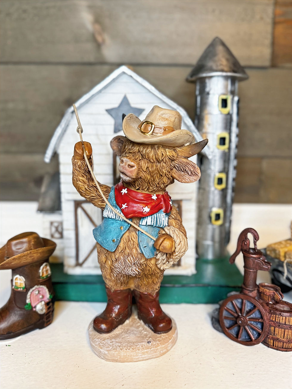 A Highland cow figurine dressed as a cowboy, wearing a hat and bandana, holding a lasso, with a barn in the background.