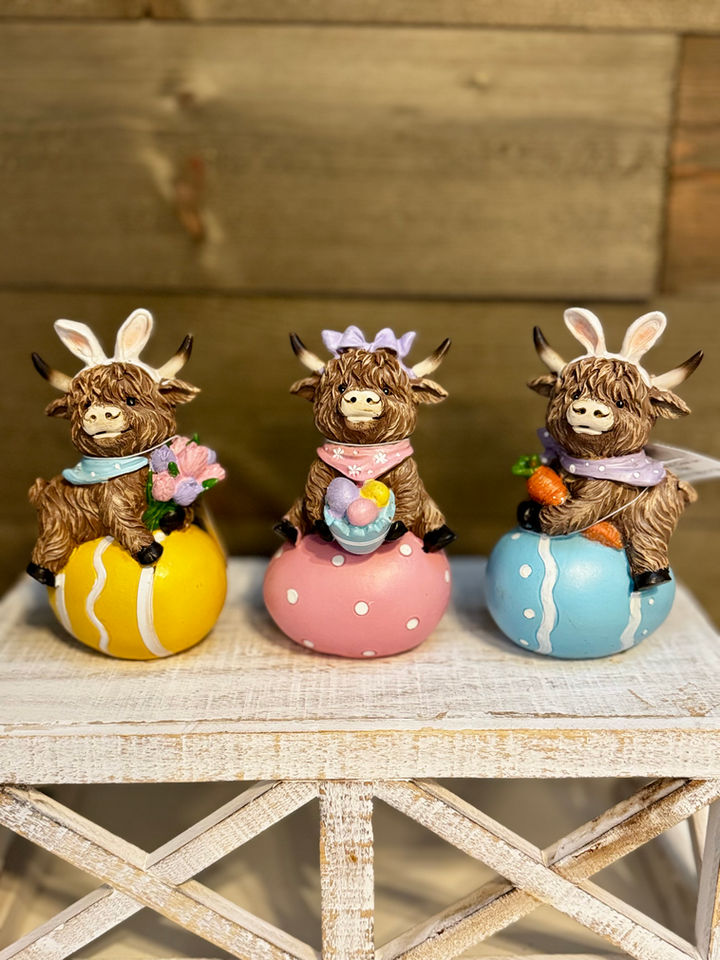 Three Highland cow Easter figurines on pastel eggs with spring flowers and carrots, charming farmhouse décor for shelves.