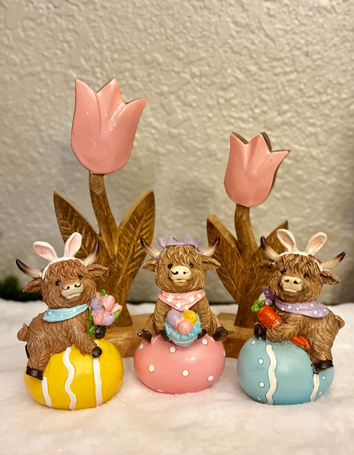 Three adorable Highland cow figurines sitting on pastel Easter eggs, wearing spring accessories, perfect for farmhouse Easter