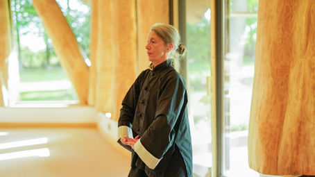 eine Frau in Tai Chi Kleidung in einer aufrechten Körperhaltung, die Arme finden in einem Kreis vor der Körpermitte zueinander. Anke Bronner im Dragonwood Tai Chi Schule Stralsund.