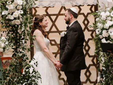 Jewish wedding ceremony at Temple Emanuel in Greensboro, North Carolina, featuring bride and groom holding hands beneath a floral chuppah with white roses and greenery, formal sanctuary backdrop, and elegant indoor wedding décor.