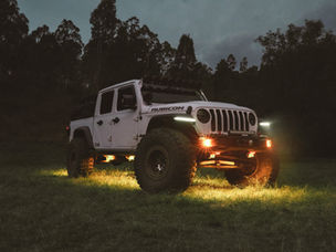 A white Jeep Gladiator Rubicon fitted with rock lights, and roof-mounted accessories, parked on grass at dusk with its lights glowing against a backdrop of trees.