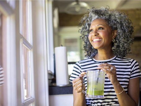 A middle-aged woman smiles while looking out a window and stirring her drink