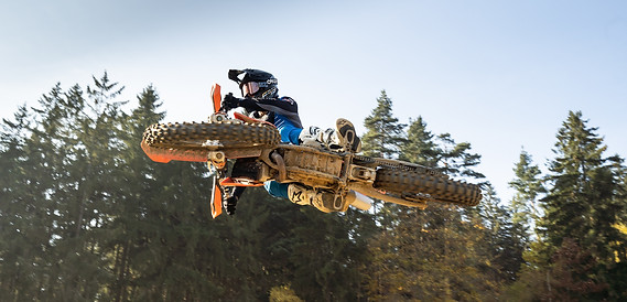 Motokrosový jezdec ve vzduchu během skoku, obklopený prachem a lesním pozadím. 