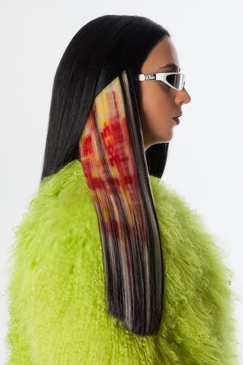 Photo of woman with long brown hair with screen printed red pattern on one bleached section. Wearing white dior sunglasses and a fluffy green coat.