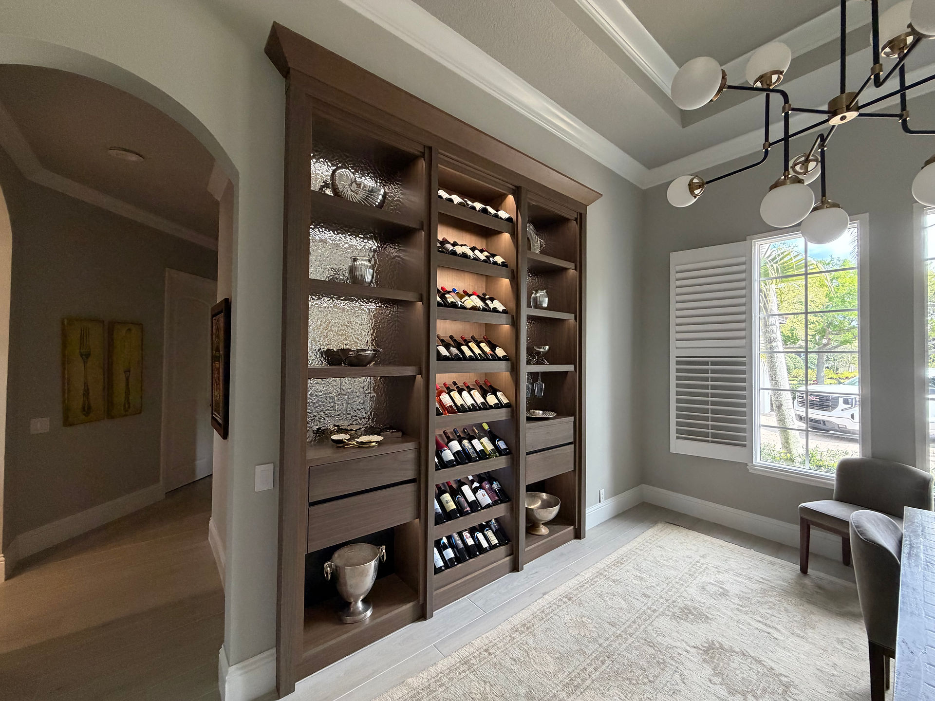 Luxury custom-built wine storage gallery in Lakewood Ranch, FL. This floor-to-ceiling unit features furniture-grade inset cabinetry, a textured hammered-metal back panel, and tiered label-forward bottle displays