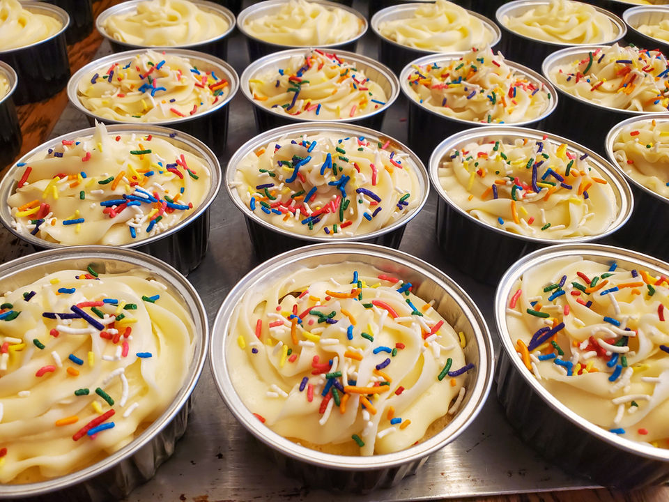 Rows of vanilla cupcakes topped with creamy frosting and colorful rainbow sprinkles, freshly made by Give A Cake Bakery volunteers for .