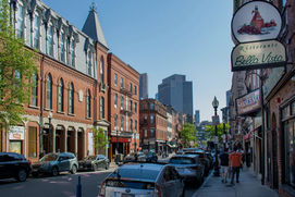 Street view of Boston's historic North End, showcasing charming red-brick buildings, Italian restaurants with iconic signs, and a vibrant atmosphere under a clear blue sky.
