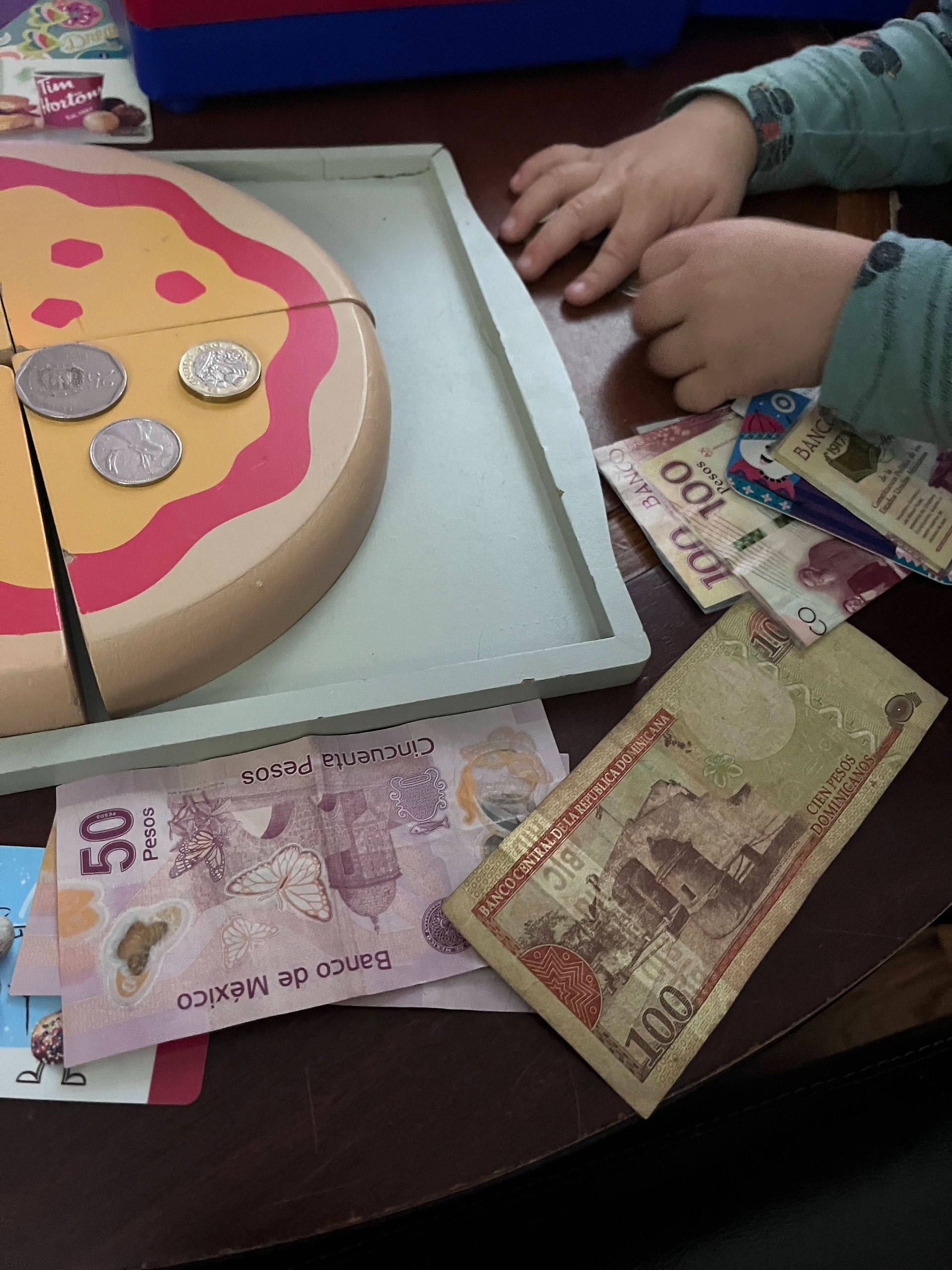 Kids hand counting coins alongside foreign currency bills and a wooden pizza set.