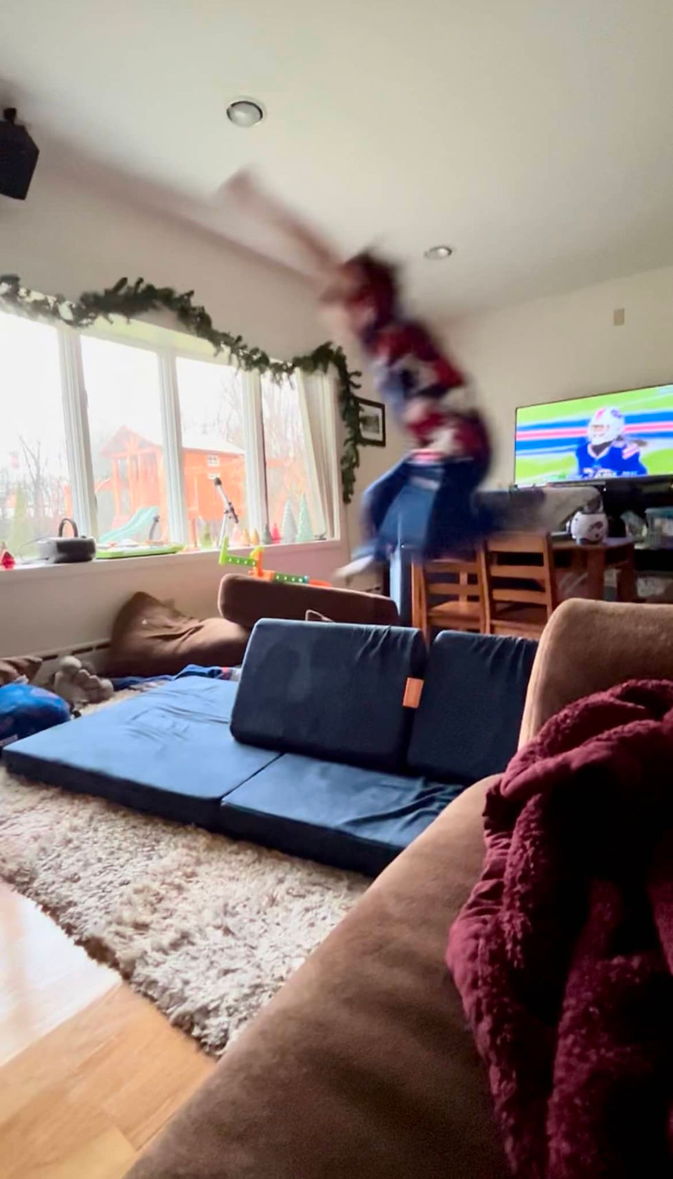 A child in motion, mid-jump, leaping over a laid out play couch in a cozy living room with a TV and windowed background.