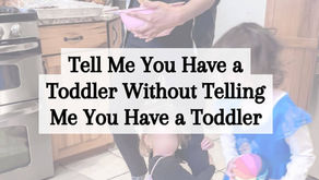 A woman mixes pancake batter in a pink bowl while two toddlers move around her in a kitchen. One toddler clings to her leg and the other wears a blue dress and holds a colorful toy. Text overlay reads, “Tell Me You Have a Toddler Without Telling Me You Have a Toddler.” Website link reads, makemindfulmemories.com.