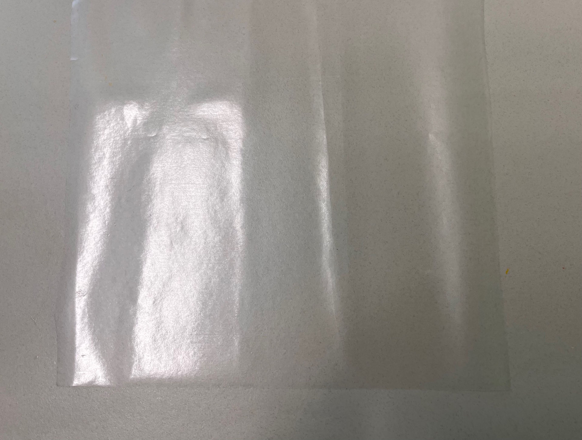 A clear plastic sheet of contact paper lies flat on a white table, casting soft light shadows, suggesting it is being used for a craft. 