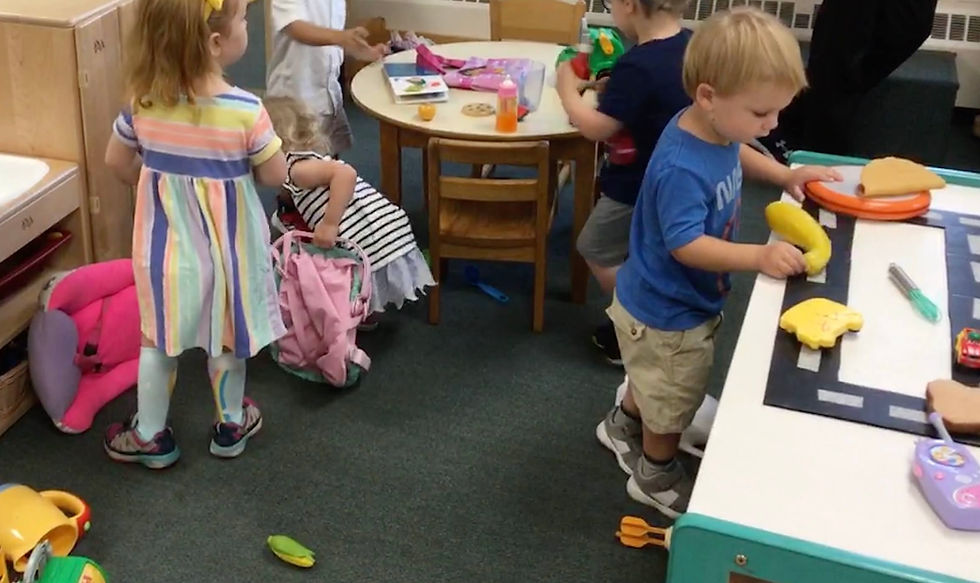 Several children are engaged in different activities in the same room, demonstrating parallel play. One child is playing with toy food items at a table, while another child is nearby playing with different toys, each focused on their own play.
