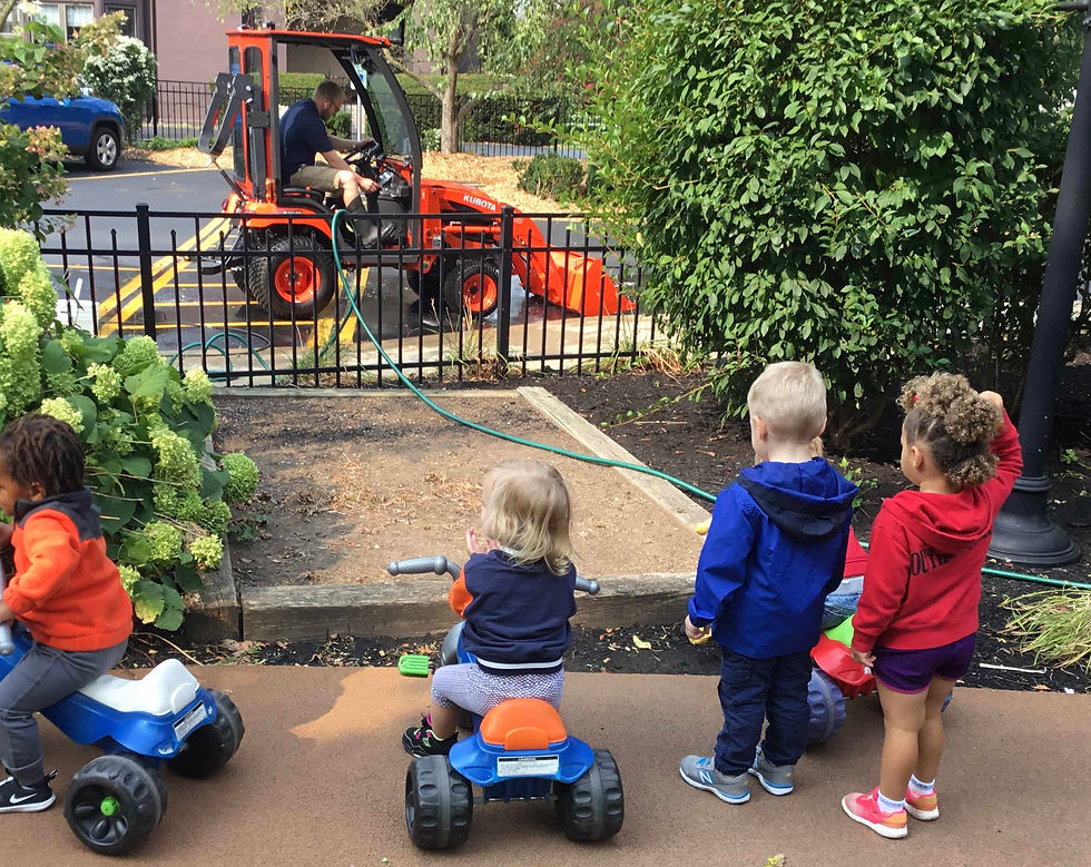 Four children are demonstrating onlooker play as they stand outside, observing a man operating a small tractor behind a fence. Two children are on tricycles, while the other two stand and watch, showing interest but not engaging.