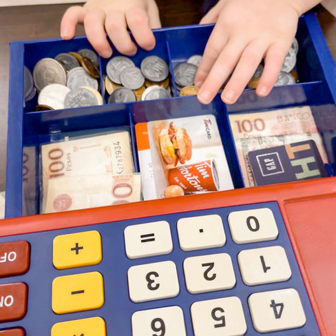 Kids' hands are opening up a pretend cash register. The register is full of foreign currency and old gift cards.