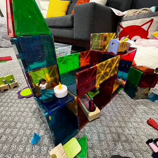 A colorful Magna-Tile structure styled like a playhouse, featuring small Encanto figurines, wooden ramps, and LED tealights, with a couch and pillows in the background.