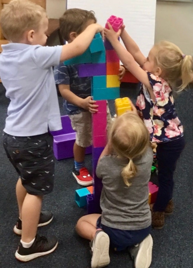 Four children are collaborating to build a tall structure using large colorful plastic blocks. They are all focused on adding pieces to the structure together, indicating cooperative play.