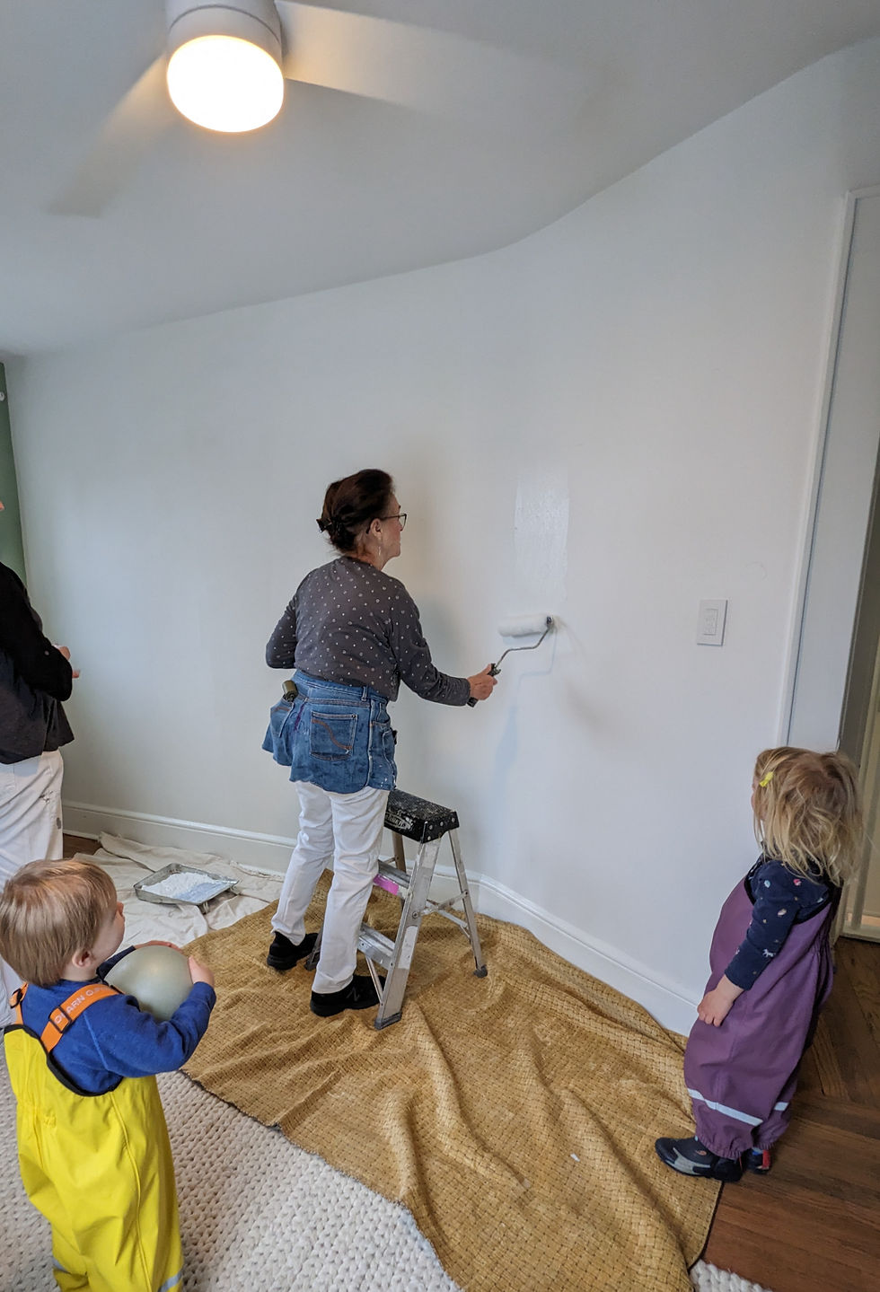 Woman prepares a white wall with roller for wallpaper installation. Ladder, drop cloths ready for wallpaper hanging.