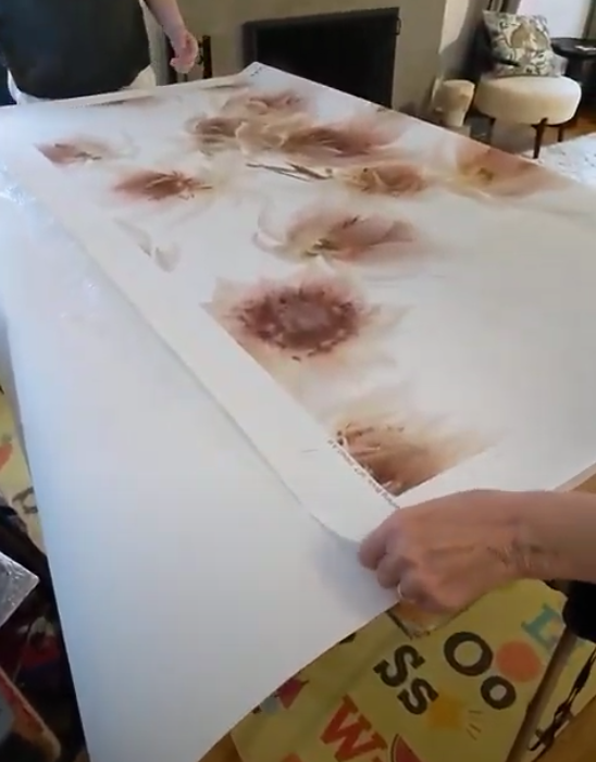 Two people roll a large floral wallpaper print on a table illustrating how to book wallpaper for hanging.