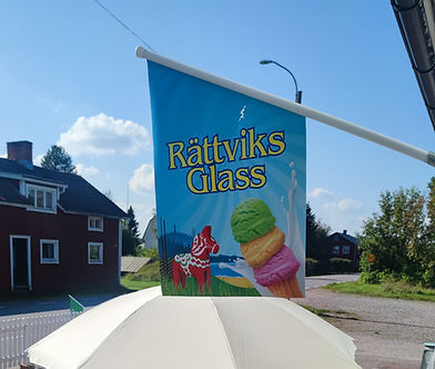 Bild på en utomhusparaply och en flagga som visar 'Rättviks Glass', med en bakgrund av en röd stuga och ett gatuområde. Det finns ett litet bord med en glasskartong på bänken och solsken.