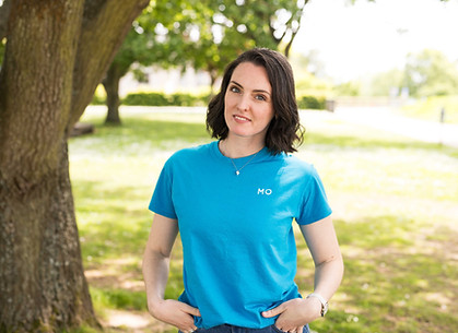 Mo wears a blue T-shirt with her name on, smiling, standing under a tree with dappled sunlight