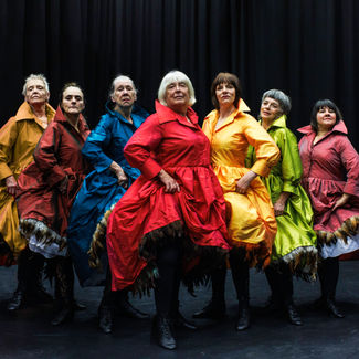 Busting Trifles. Moving Memory Theatre Company. Seven mature light skinned women stand proudly in a line, holding the puffy skirts of their bright silky dresses.