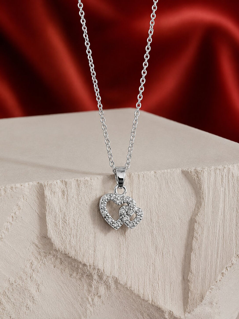 Double Heart Silver Necklace with sparkling crystal heart pendant displayed on a textured stone platform with elegant red sat