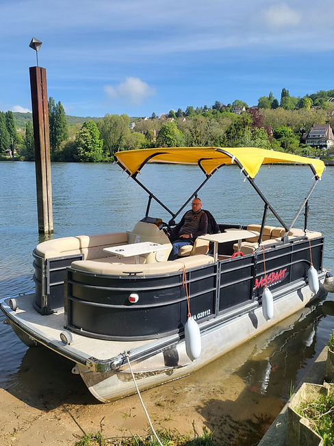 Une photographie prise en plein jour montre un ponton de luxe, "Évasion Nautique - Triel-sur-Seine", amarré au bord d'une rivière en France. Un homme portant des lunettes et une veste sans manches est assis au poste de pilotage, face à l'arrière. Le ponton est noir et gris, avec des sièges en cuir beige et un auvent jaune vif déployé. Deux petites tables blanches sont installées à bord. Deux pare-battages blancs pendent sur le côté. En arrière-plan, la rivière s'étend sous un ciel bleu parsemé de quelques nuages, avec une rive boisée et des collines habitées. Un grand poteau en bois surmonté d'un réflecteur radar est visible sur la gauche