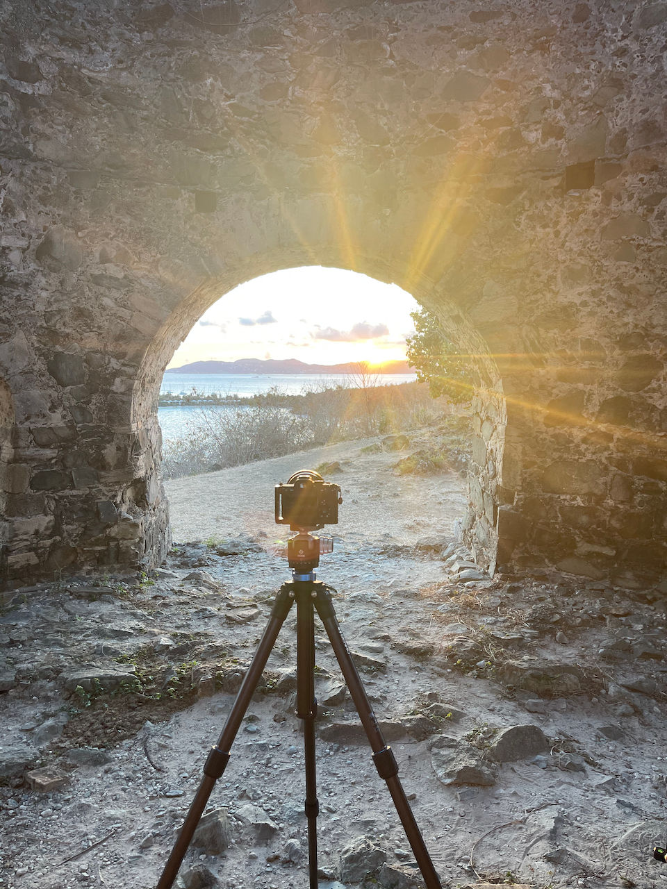 the sun bursts from the horizon as a camera mounted on a tripod peers through a stone arch to capture the beauty of a rocky shoreline, the ocean, and the mountains on the other side of the Bay