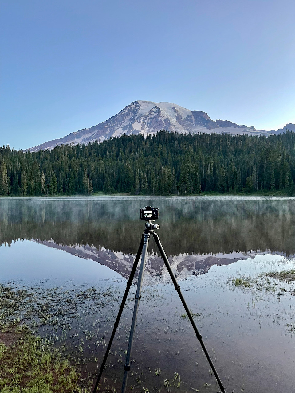 A camera mounted on a tripod at the shore of a crystal clear reflective lake, photographing the glory of the enormous mountain rising above the trees on the opposite shore - and the reflection of all of this in the water below