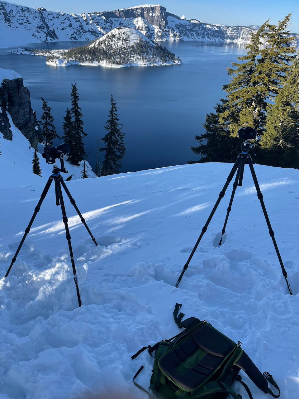Snow covers Wizard Island and the shore of Crater Lake in Oregon. Two cameras are mounted on tripods on the shore to capture the majesty
