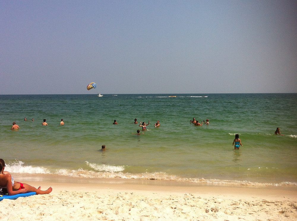 the sandy shores of the Gulf of Mexico in Southern Alabama. Beachgoers enjoy the warm water and watch the parasailer in the background