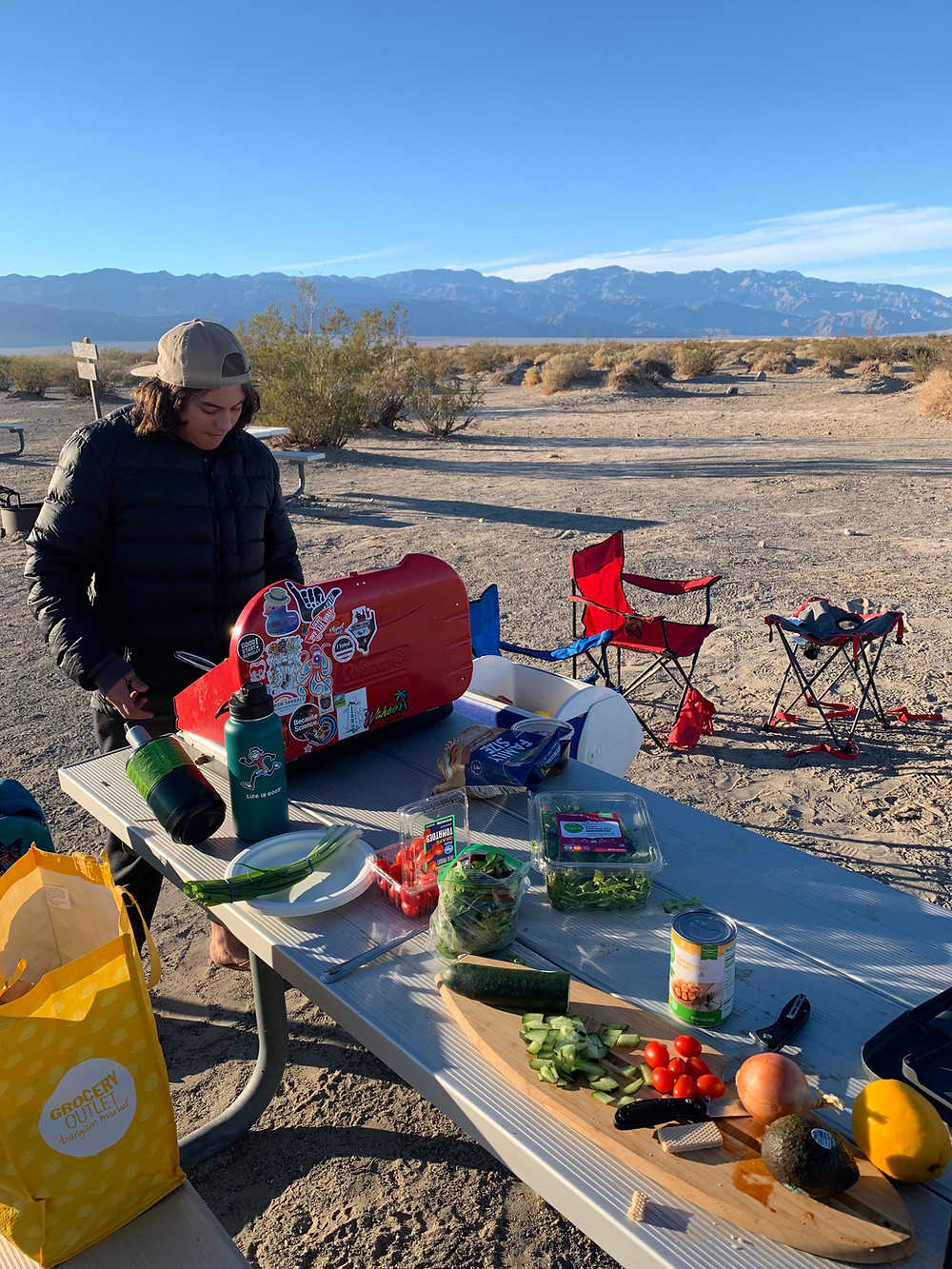 A woman wearing black and a backwards hat cooks at her campsite, all her cooking gear and groceries laid out on a white table in the middle of the desert