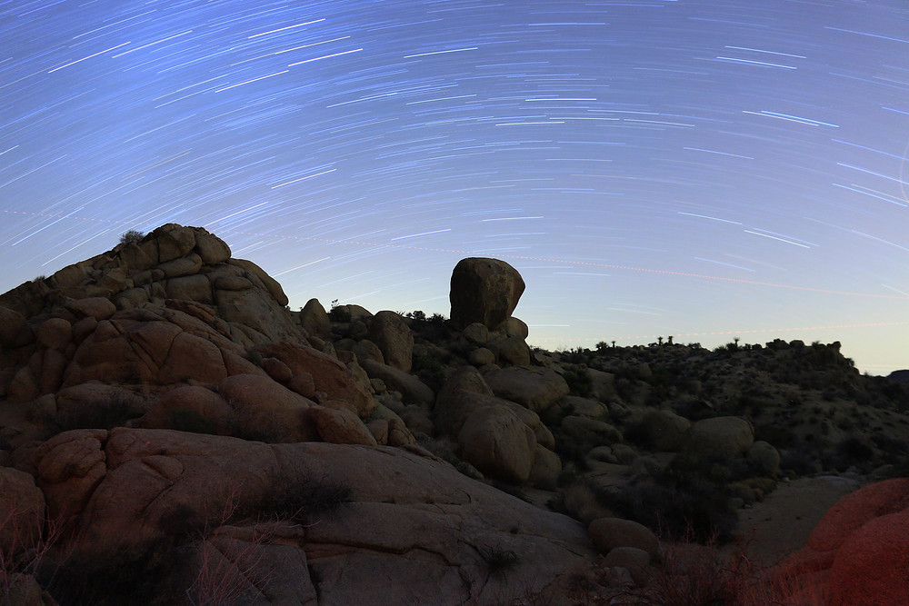 a timelapse photo of the starscape above the dark rocky craggy outlines of Joshua Tree National Park