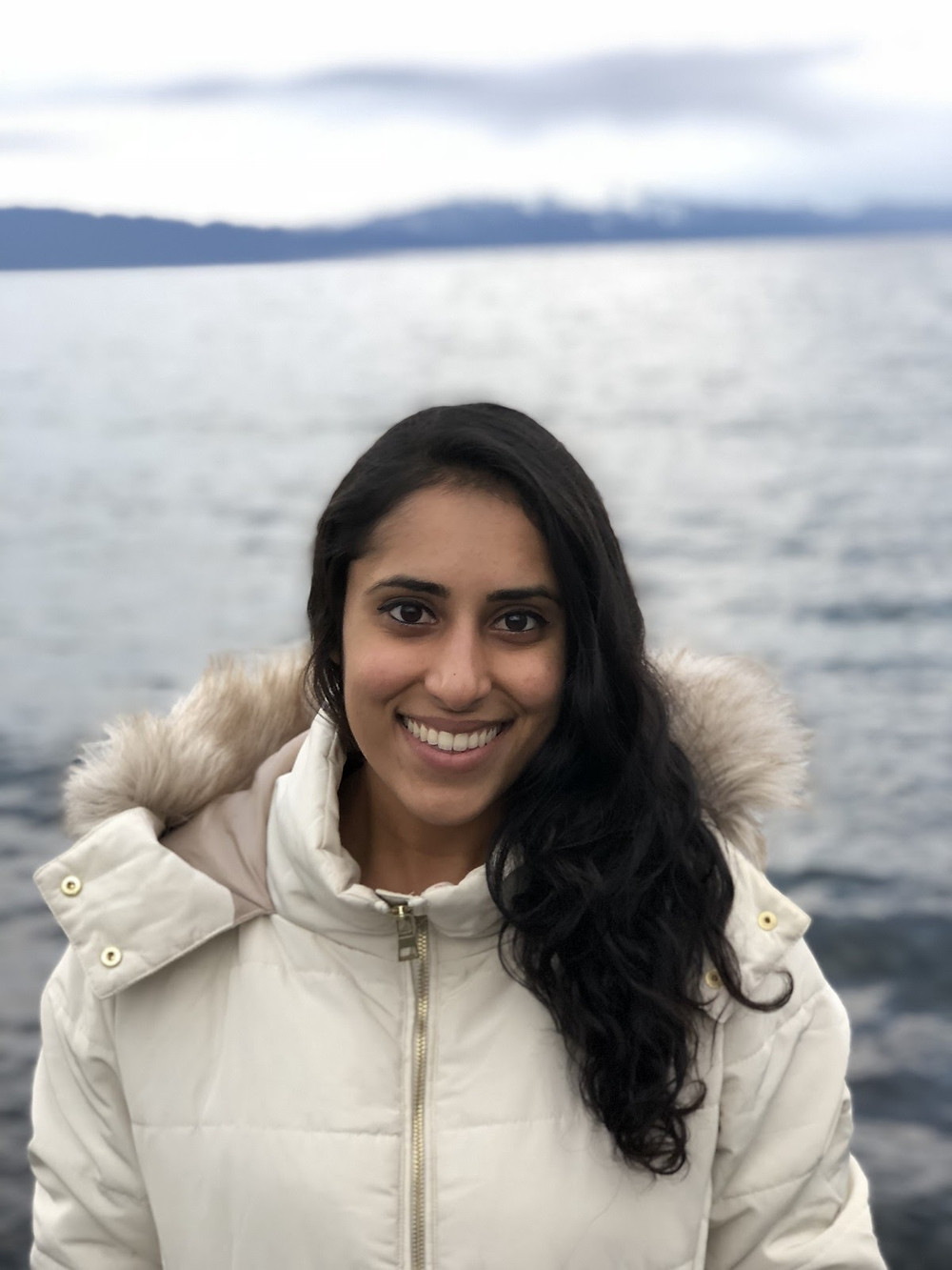Guest blogger Ashima smiles for the camera wearing a white parka in front of a beautiful body of water