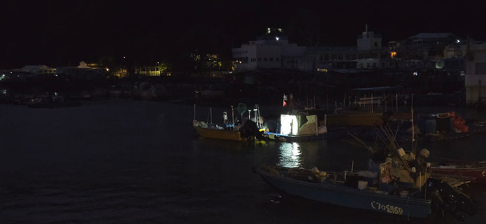 Boats on a dark harbor at night, one brightly lit. Buildings visible in the background, with a calm and quiet atmosphere.