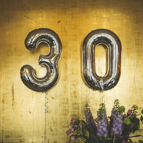 Silver "30" balloons against a gold background with purple flowers below. Celebratory and festive atmosphere.