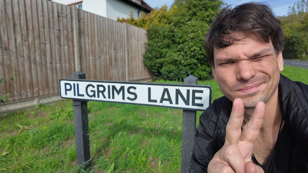 Man takes a selfie beside a Pilgrims Lane street sign, squinting and flashing a peace sign on a sunny suburban roadside.