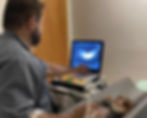 Veterinário examina cãozinho em ultrassom. Sala com paredes claras e porta de madeira. Monitor exibe imagem médica.