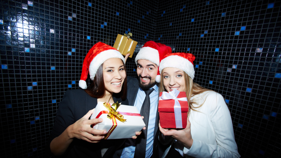 A male and two female colleagues wearing Santa hats and holding up presents at their work Christmas party.