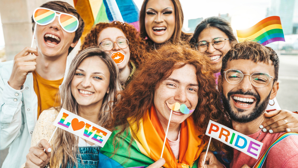 A diverse group of young people, emiling and holding up rainbow and Pride flags.