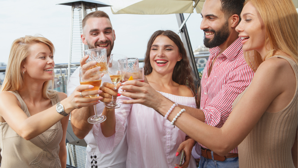A mixed group of young professionals, smiling and clinking glasses in a toast, at their company summer party at one of the top London rooftop venues for summer events.