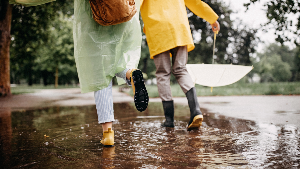 A rear view of a woman in a white raincoat, cropped blue and white striped trousers and yellow ankle wellies, arn in arm with a man in a yellow mac and grey checked trousers ticked int wellies, splashing through a large puddle.