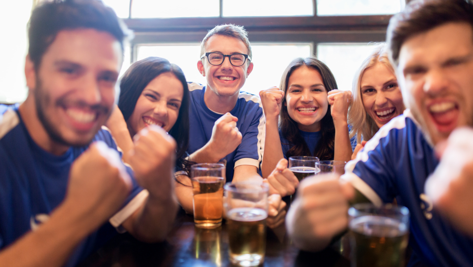 A group of young professional men and women in blue football shirts, punching the air and smiling, sat a a table with drinks of beer, at World Cup corporate events UK.