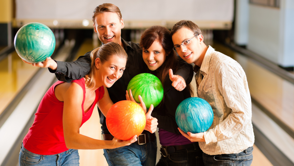 A group of young colleagues holding different coloured bowling balls at a 10 pin bowling alley.