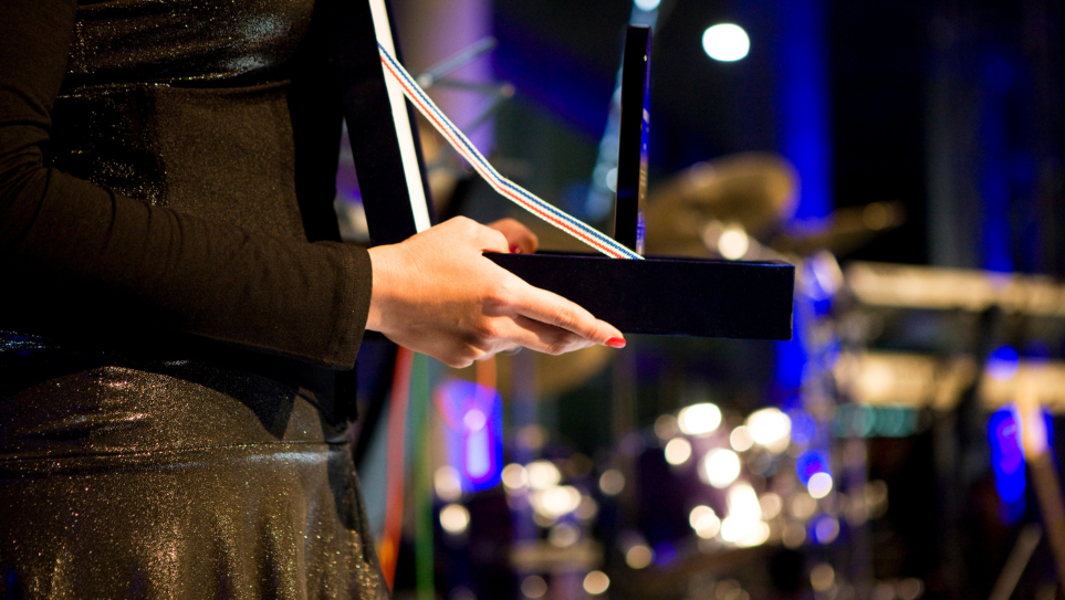 A woman in a black and gold dress holding an open box with a company award at a company awards gala.