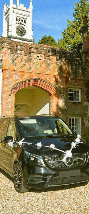 Black Mercedes-Benz V-Class decorated with white wedding ribbons parked in front of a brick estate archway of the Farnham Castle.