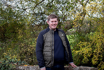 Smiling man in a jacket standing by a stone wall outdoors.
