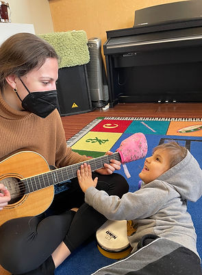 A young brunette woman is sitting cross-legged on a colorful rug. She is playing the guitar. The woman is wearing a brown turtle neck sweater and black pants. A young boy with blond hair is lying on the carpet in front of the woman, and he is reaching out with his left hand and touching the fretboard of the guitar. The boy is in a gray hoodie sweatshift with the hood up, and a pair of dark gray sweat pants with a light gray and white stripe on the side. The boy is smiling as if he's enjoying the interaction he's having with the woman.