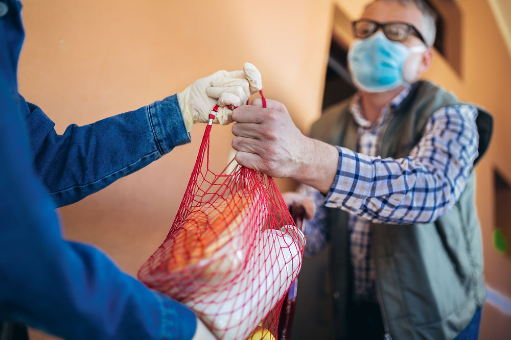 Person wearing gloves hands bag of groceries to person wearing mask.